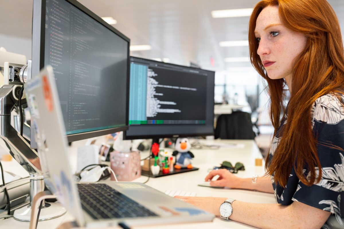 woman software developer with red hair looking at lines of computer code at a desk