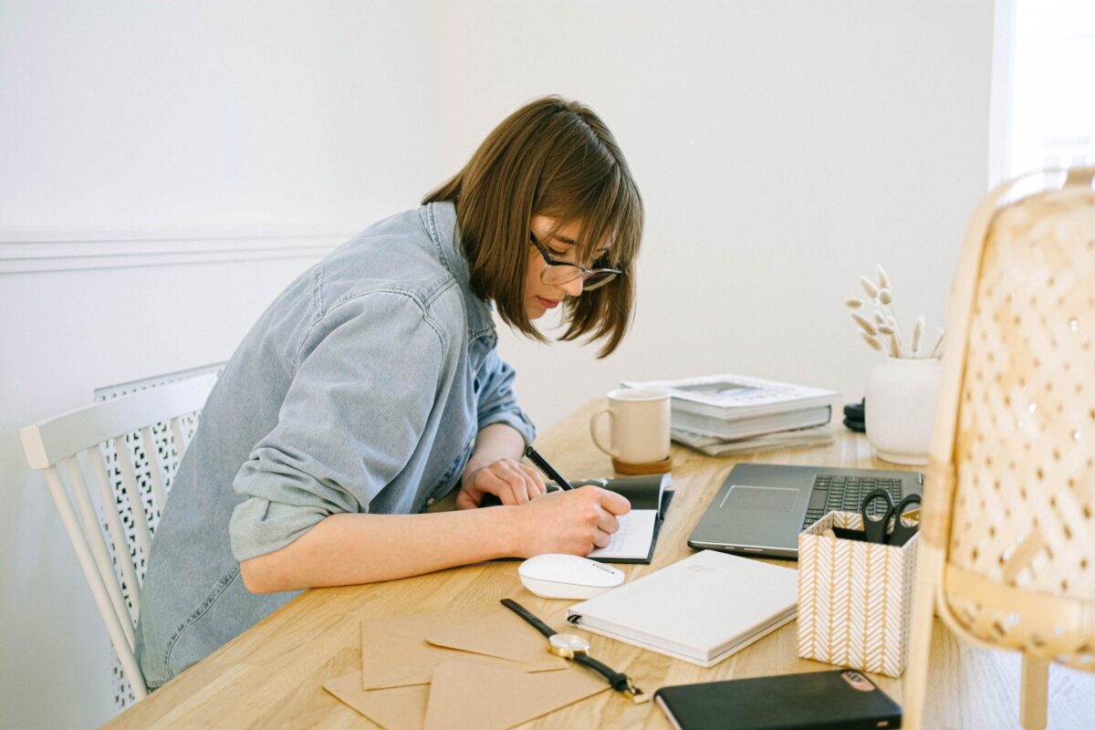 a woman writing in a journal at a desk with a computer in front of her