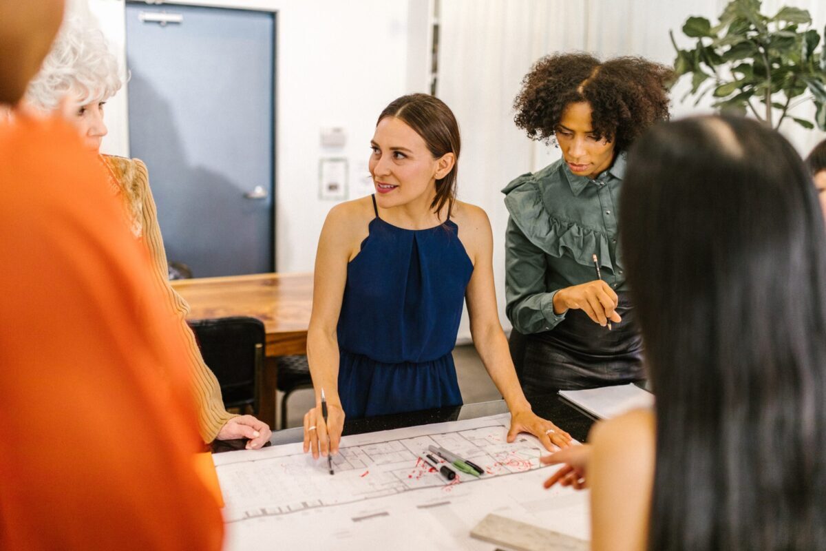 a woman standing at the head of a table with multiple other people surrounding her